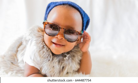 Super cool baby. Hipster baby in fur vest and sunglasses lies on a white bed in a room with curtains. Baby smiles while putting on sunglasses. 