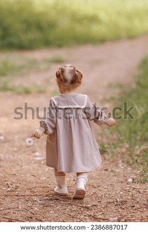 Super cute anonymous blonde with curly hair in a white dress walking along a rural road. Small child girl walking in the park. Rear view.