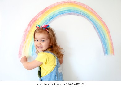 Super cute little girl drawing rainbow on white wall at home. Sign of hope. Quarantine. Coronavirus. Smiling child looking in camera. Stay at home.