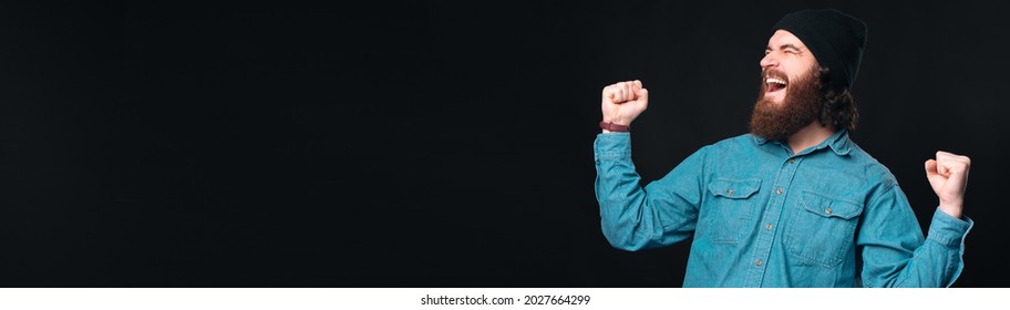 Super Excited Bearded Man Is Making The Winner Gesture Over Black Background. Banner Sized Photo.