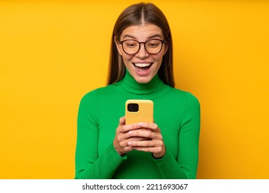 Super Excited Overjoyed Young Woman In Stylish Glasses And Green Clothes Holding Smartphone With Surprised And Excited Face Expression After Receiving Message On Yellow Background