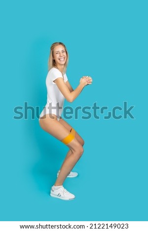 Super excited woman doing squats with orange resistance band on legs smiling with teeth in white bodysuit on blue background. Doing sports to be in good shape. Improving health by yourself at home.