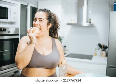 Super happy plus size curly woman smiling to camera while eating fruit at the kitchen home to lose weight. Training clothes. Getting fit for the summer concept.Beach body preparation, healthy life