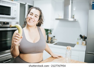 Super happy plus size curly woman smiling to camera while eating fruit at the kitchen home to lose weight. Training clothes. Getting fit for the summer concept.Beach body preparation, healthy life