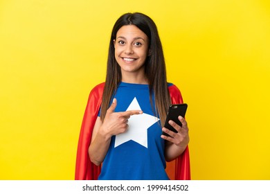 Super Hero woman isolated on yellow background using mobile phone and pointing it