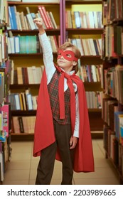 Super Kid. A Boy Dressed In School Uniform And Superhero Red Cape And Mask Stands In A Library By Bookshelves Holding One Arm Up. Super Abilities In Education. Children's Dreams. 