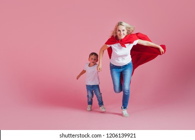 Super Mom And Her Son Run Forward. Fun Family, A Young Blond Woman In A Red Cape As Superhero, Pink Background