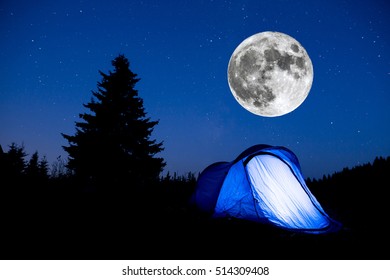 Super Moon. Blue Illuminated Tent With Travelers In The Mountain. Background Of A Pine Tree Silhouette And The Starry Summer Night Sky.