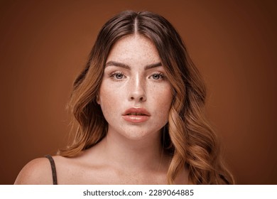 Super natural, beautiful girl looking at the camera. Attractive woman with freckles on her face and long wavy hair. Brown studio background. - Powered by Shutterstock