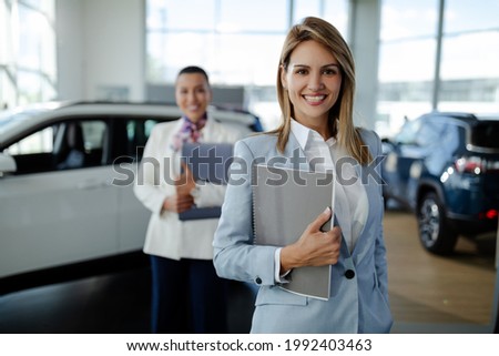 Super salesteam in dealership, two consultants or managers in elegant suit with laptop computer in arms looking on camera with smile.