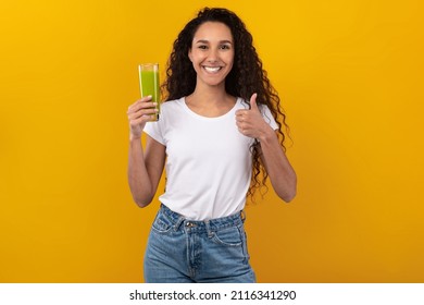 Superfood Healthy Detox And Diet Food Concept. Portrait Of Smiling Young Woman Holding Glass Of Green Smoothie Drink Showing Thumbs Up Gesture Standing Isolated On Yellow Orange Studio Background Wall