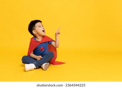 Superhero Asian boy with red cape sitting on floor and pointing to copy space isolated yellow background, Full body composition - Powered by Shutterstock