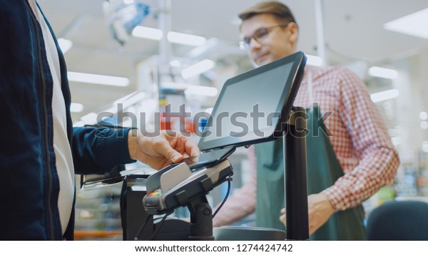 At\
the Supermarket: Checkout Counter Customer Pays with Smartphone for\
His Food Items. Big Shopping Mall with Friendly Cashier, Small\
Lines and Modern Wireless NFC Paying Terminal\
System.