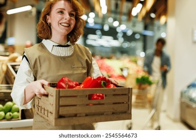 Supermarket employee carrying a wooden crate full of red bell peppers, smiling and looking away, while working in the fresh produce section of a grocery store - Powered by Shutterstock