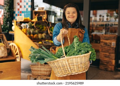 Supermarket Employee With Down Syndrome Holding A Basket Of Fresh Organic Vegetables In A Grocery Store. Happy Woman With An Intellectual Disability Working In A Local Convenience Store.