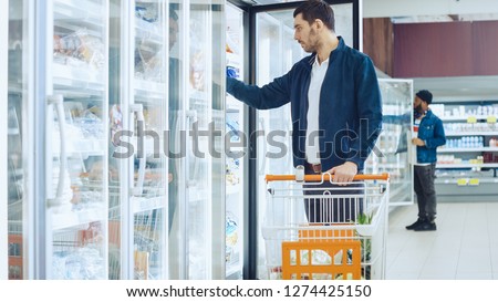 At the Supermarket: Handsome Man Pushes Shopping Card and Browses for Products in the Frozen Goods Section. Man Opens the Fridge Door and Takes Frozen Vegetables. Other Customer Shopping in Background