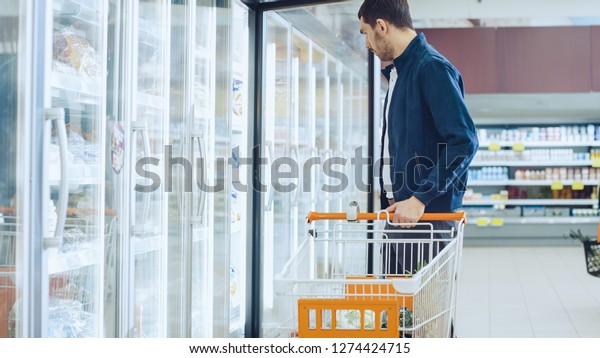 At the Supermarket: Handsome Man Pushes\
Shopping Card and Browses for Products in the Frozen Goods Section.\
Man Opens the Fridge Door, Looking for Dairy Products. Other\
Customer Shopping in\
Background