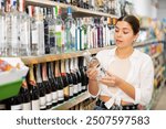 in supermarket in hard liquor department, young woman carefully chooses vodka