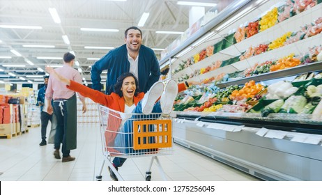 At The Supermarket: Man Pushes Shopping Cart With Woman Sitting In It, Happy Couple Has Fun Racing In A Trolley Through The Fresh Produce Section Of The Store.