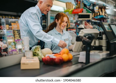 Supermarket Manager Training Young Worker At Checkout Counter. Senior Man Teaching The Checkout Process To New Employee In Grocery Store.