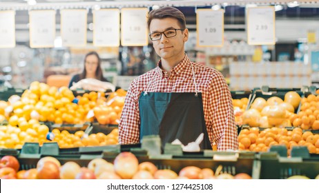 At The Supermarket: Portrait Of The Handsome Stock Clerk Wearing Apron, Arranging Organic Fruits And Vegetables, He Smiles Into Camera. Friendly, Efficient Worker At The Store.