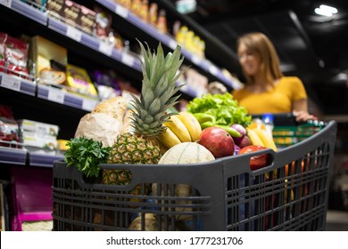 Supermarket Shopping. Close Up View Of Shopping Cart Overloaded With Food While In Background Female Person Choosing Products.