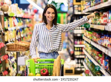 At The Supermarket. Smiling Young Woman Standing With Trolley Cart Between Aisles In Grocery Store. Cheerful Consumer Buying Essentials In Super Market, Taking Products From Shelf Looking At Camera