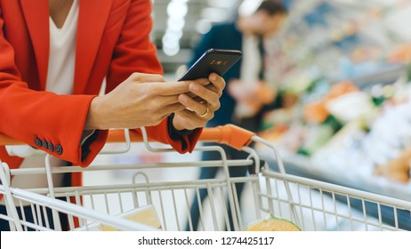 At the Supermarket: Woman Uses Smartphone, Leans on the Shopping Cart. In the Big Mall Woman Browsing In Internet on Her Mobile Phone. Focus on Hands Holding Mobile Phone. - Powered by Shutterstock