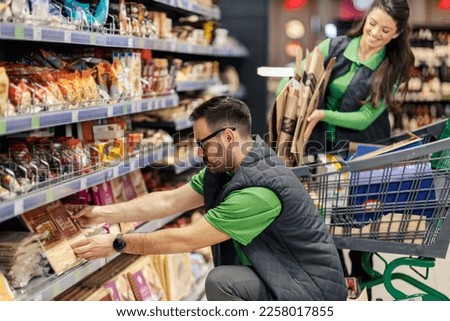 A supermarket worker is crouching next to an aisle and putting products and groceries on shelf.