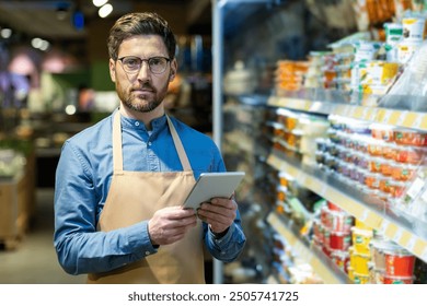 Supermarket worker standing in grocery aisle with tablet, dressed in apron and glasses. Busy retail environment with various food products on shelves - Powered by Shutterstock