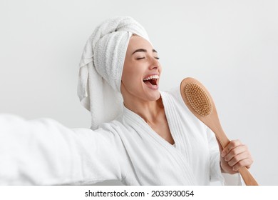 Superstar In Bathroom, Enjoy Morning Treatments At Home, Happy Routine. Funny Smiling Beautiful Young Caucasian Woman In Robe And Towel On Head Singing In Brush On White Wall Background, Empty Space