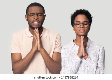 Superstitious ethnic millennial couple wearing glasses isolated on grey studio background hold hands in prayer, hopeful black biracial man and woman believer pray make wish hope for best