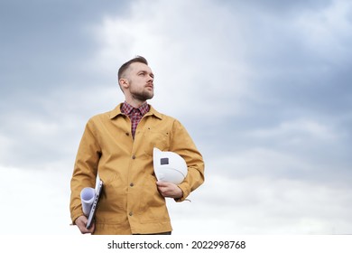 Supervising on site concept. Serious cute caucasian bearded construction contractor, supervisor, foreman or architect with blueprints, laptop and helmet in hands at construction site in orange jacket
