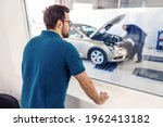 The supervisor oversees the work in the car service. A man in a blue T-shirt stands in the office looking out the window of a worker performing a technical inspection of a car with a raised hood