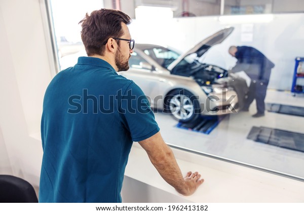 The supervisor oversees the work in the car service.\
A man in a blue T-shirt stands in the office looking out the window\
of a worker performing a technical inspection of a car with a\
raised hood