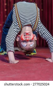 Supple Agile Little Boy Doing A Back Arch Handstand On Stage In A Theatre Wearing His Colorful Makeup As He Grins At The Camera From His Upside Down Position