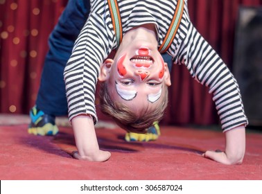 Supple Agile Little Boy Doing A Back Arch Handstand On Stage In A Theatre Wearing His Colorful Makeup As He Grins At The Camera From His Upside Down Position