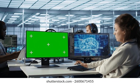IT support workers in AI data center coding on green screen computers, upgrading equipment. Multiethnic team of engineers at PC desk overseeing server clusters using mockup monitors, camera B