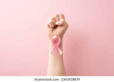 Supporting Breast Cancer awareness concept. First-person perspective of strong raised hand with clenched fist. Pink ribbon attached to wrist showcased on pastel pink surface with room for text