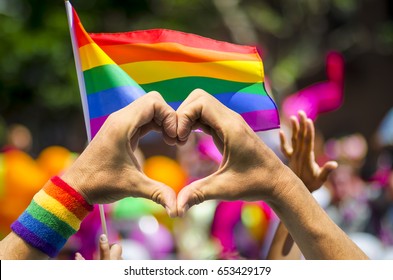Supporting hands make heart sign and wave in front of a rainbow flag flying on the sidelines of a summer gay pride parade - Shutterstock ID 653429179