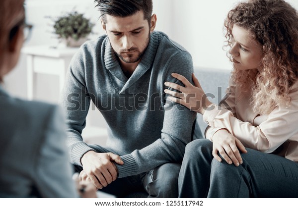 Supportive beautiful\
wife touching husband\'s arm during psychotherapy session for\
married couples with\
problems