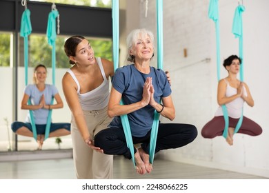 Supportive female trainer helping to mature female balancing in Lotus Pose, performing antigravity aerial yoga exercise in studio - Powered by Shutterstock