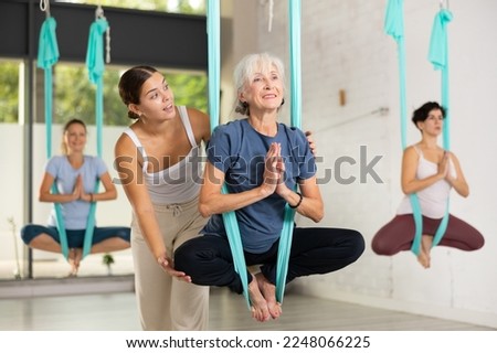Supportive female trainer helping to mature female balancing in Lotus Pose, performing antigravity aerial yoga exercise in studio