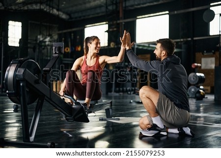 Supportive fitness instructor giving high-five to athletic woman after exercising on rowing machine during cross training in a gym. 