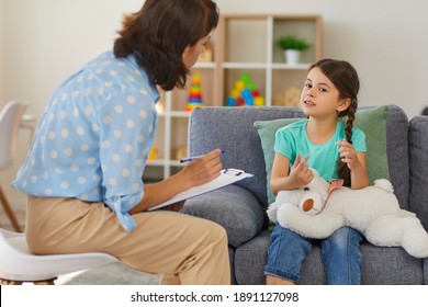 Supportive Psychologist With Clipboard Listening To Little Child During Therapy Session. Preschool Girl Feeling At Ease In Therapist's Office Sharing Her Thoughts And Concerns