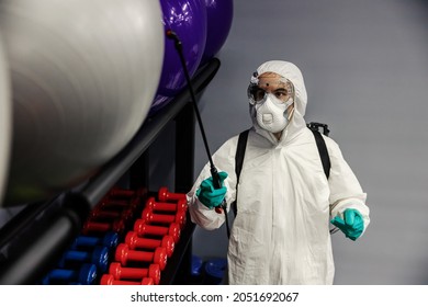 Suppression Of COVID-19 Virus And Cleaning Of Exercise Equipment In The Gym By A Male Person In A Protective White Suit. The Focus Of The Photo Is On The Pilates And Yoga Balls