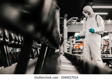 Suppression Of COVID-19 Virus And Cleaning Of Neatly Arranged Exercise Equipment In The Gym By A Male Person In A Protective White Suit.Antiseptic, Disinfection, Cleanliness, Healthcare, Antibacterial