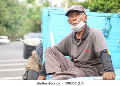 Surabaya, Indonesia - May 23, 2021: A Garbage Man Uses A Blue Garbage Cart And Is Resting Sitting On His Cart, Wearing A Long-sleeved T-shirt, Mask And Hat.