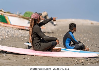 Surf instructor with dreadlocks pointing, teaching a young student surfing techniques on a sandy beach with surfboards