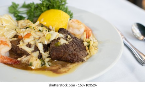 A Surf And Turf Entree Topped With Crab Meat And A Seafood Sauce Plated And Garnished With A Lemon And Kale, On A White Linen Tablecloth.